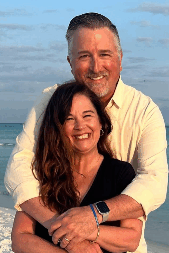 Best friend and owner, Kris standing with her husband standing on the beach smiling.